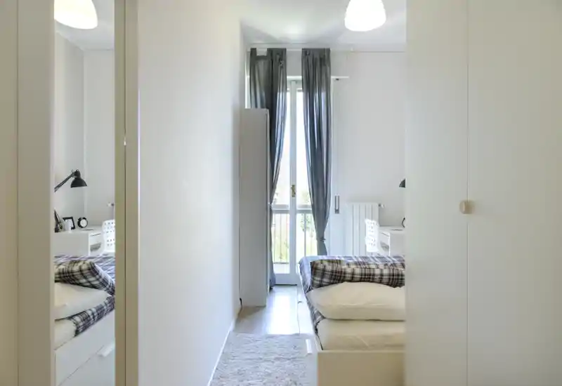 Bright, minimalist bedroom with a single bed dressed in patterned bedding, a small desk and a balcony window that brings abundant natural light.