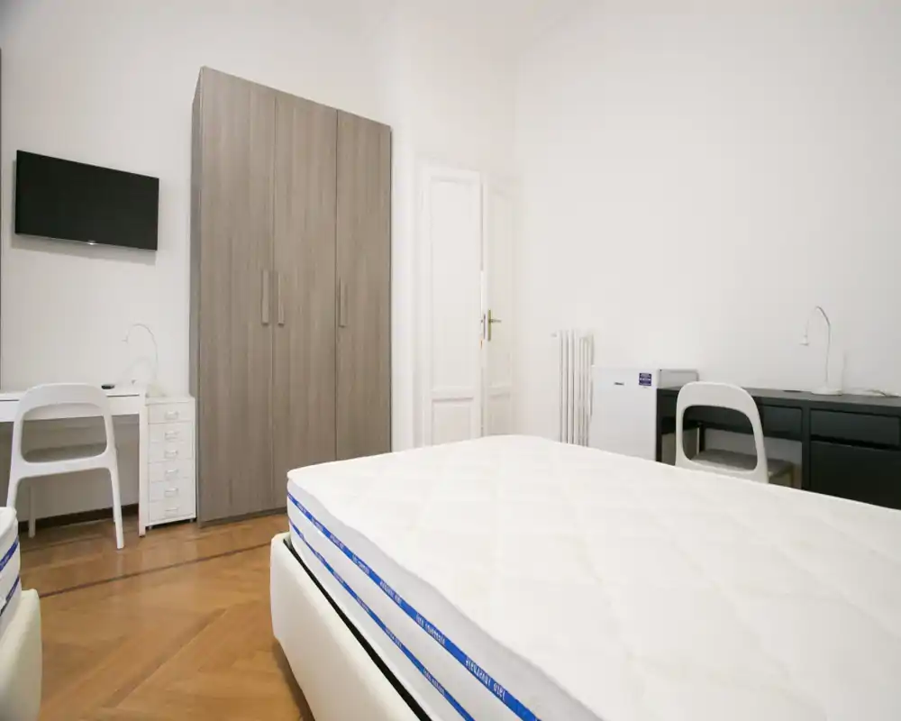 Dormitorio luminoso y minimalista con colchón visible, suelo de parquet, armario y dos escritorios — ordenado y listo para huéspedes.