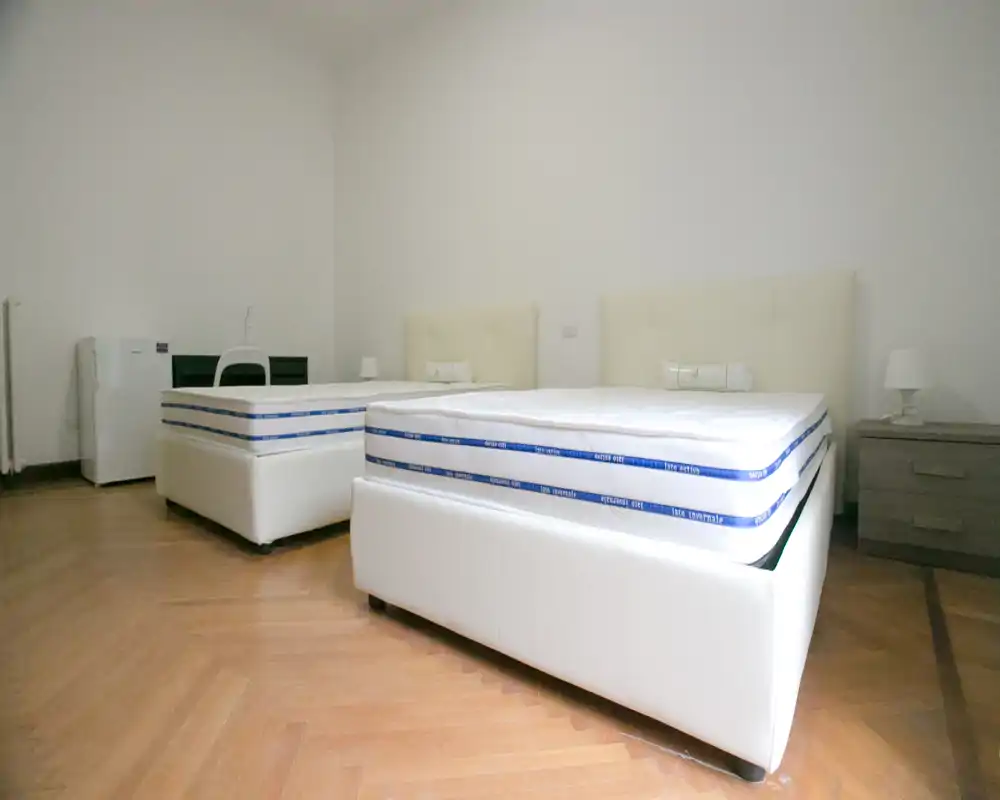 Dormitorio moderno y luminoso con dos camas tapizadas, cabeceros neutros y mesitas; suelo de parquet limpio y decoración mínima para un ambiente despejado.