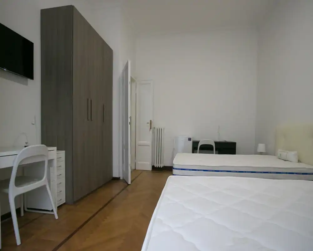 Amplia habitación amueblada de forma sencilla con dos colchones individuales, lámpara de noche, armario y escritorio — habitación luminosa con suelo de parquet y decoración neutra.