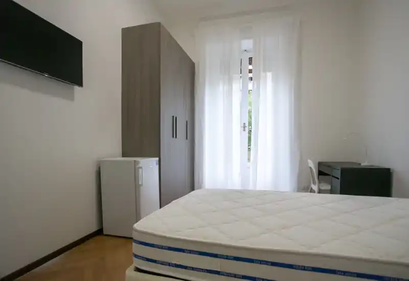 Dormitorio luminoso y de mobiliario sencillo con colchón visible en primer plano, armario, escritorio y TV mural. La ventana con cortinas translúcidas aporta luz natural suave.