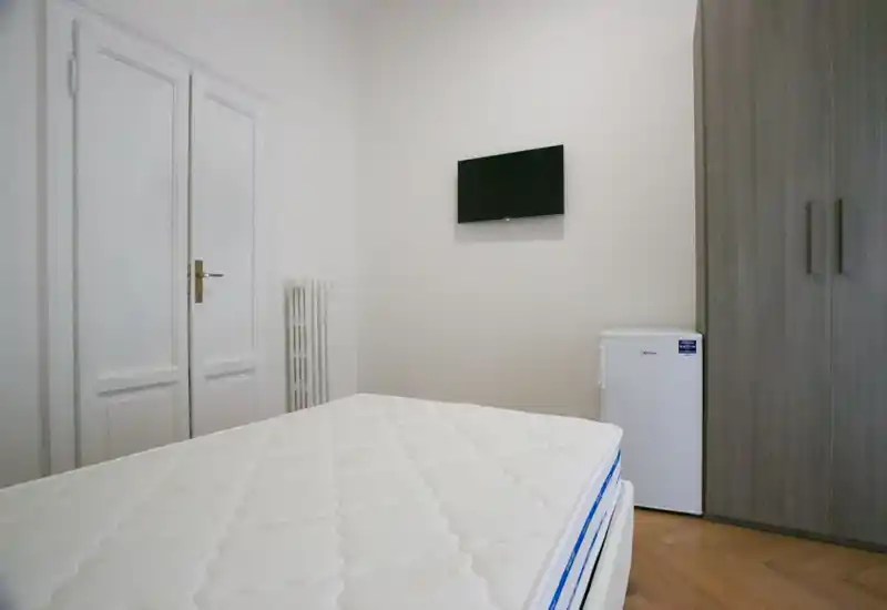 Dormitorio minimalista y luminoso con un gran colchón en primer plano, TV en la pared, armario y pequeño frigorífico; acabados modernos y neutros.