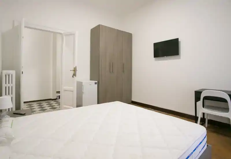 Dormitorio luminoso y de mobiliario mínimo con un colchón grande en primer plano, armario, mini nevera, TV mural y escritorio. Decoración neutra y buena iluminación, apto para alquileres de corta estancia.