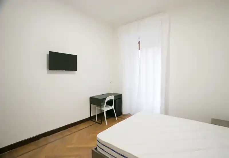 Dormitorio sencillo y luminoso con colchón visible, televisor mural y un escritorio compacto junto a una ventana con cortina traslúcida. Estilo limpio y minimalista con buena luz natural, ideal para anuncios de alquiler que destaquen la zona de descanso.
