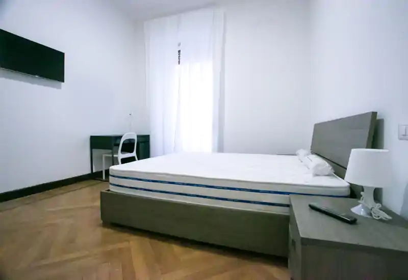 Dormitorio luminoso y minimalista con una cama grande sobre estructura de madera, mesita con lámpara y un pequeño escritorio junto a la ventana con cortina translúcida — moderno y ordenado.