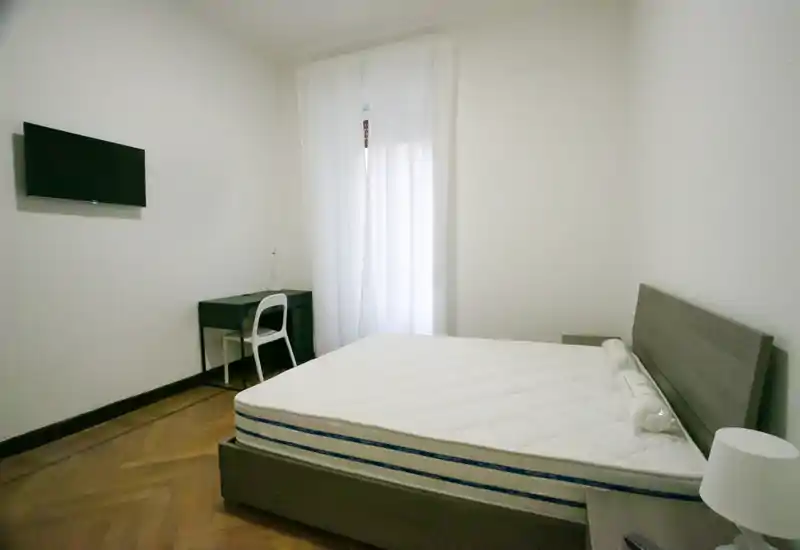 Dormitorio minimalista y luminoso con cama doble, cabecero de madera, mesita y un pequeño escritorio junto a la ventana. La luz natural se filtra a través de las cortinas translúcidas, generando un ambiente sereno y despejado ideal para alquileres.