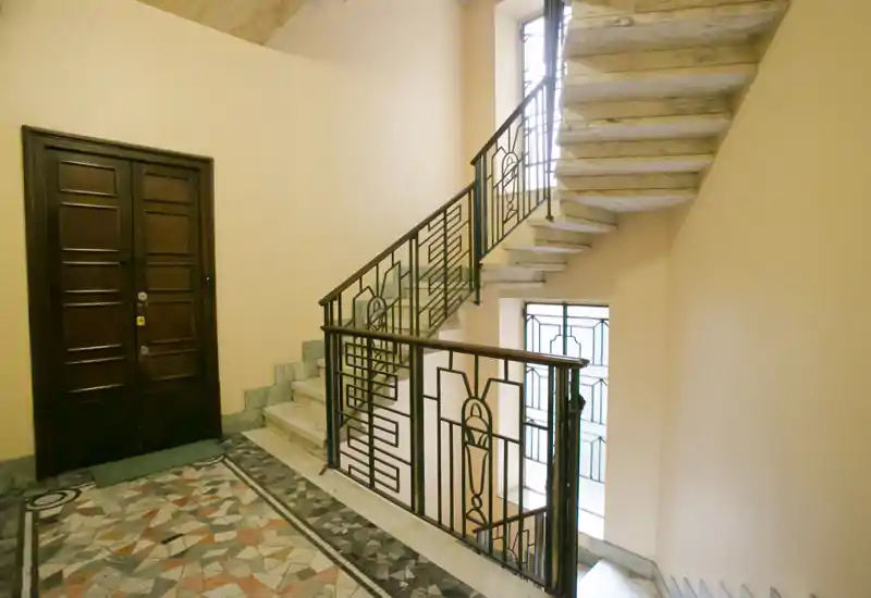 Entrada del edificio bien iluminada con suelo de mosaico decorativo, puerta de madera y barandillas de escalera de metal ornamentadas; tramo de escalera luminoso hacia los pisos superiores.