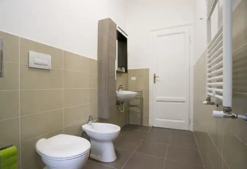 Bagno moderno e pulito con wc, bidet, lavabo a parete e colonna contenitore; piastrelle neutre e illuminazione brillante rendono lo spazio funzionale e fresco.