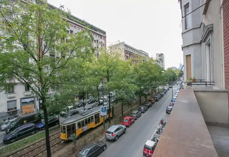 Balcone con vista su una via cittadina alberata, linea tranviaria e auto parcheggiate — utile per mostrare il contesto urbano e i trasporti.