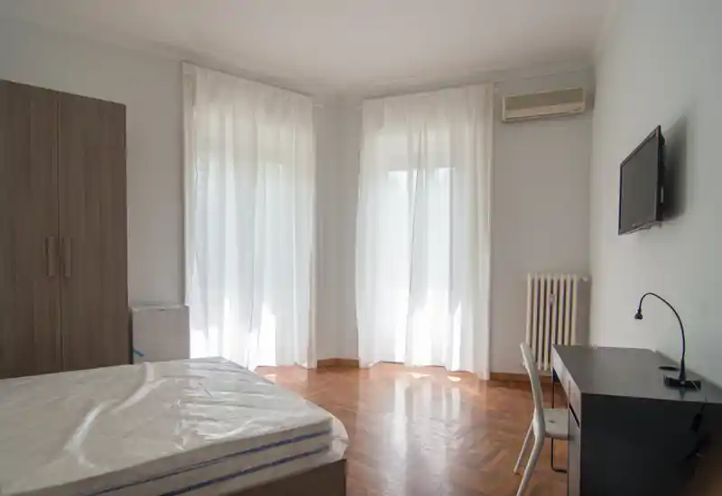 Camera da letto luminosa e minimal con materasso in vista, pavimento in parquet, armadio, scrivania e TV a muro. Le tende leggere diffondono la luce naturale rendendo l'ambiente tranquillo.