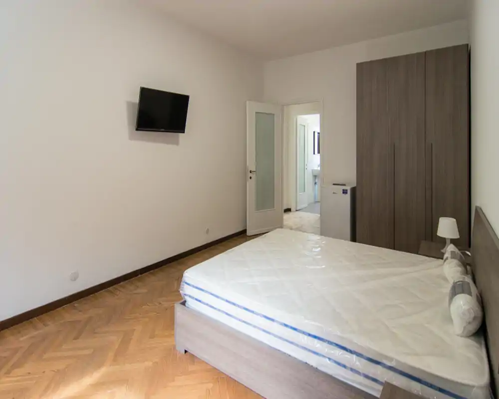 Camera da letto luminosa e arredata in modo essenziale con letto matrimoniale, armadio e TV a parete. Il materasso è ancora con pellicola protettiva, pavimento in parquet e pareti neutre.