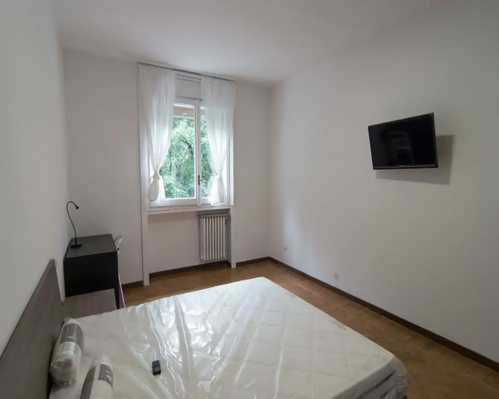 Camera da letto luminosa e semplice con materasso matrimoniale in primo piano, luce naturale da una finestra con tende leggere, comodino con lampada e TV a parete. Arredamento essenziale e pulito, adatto per annunci in affitto.