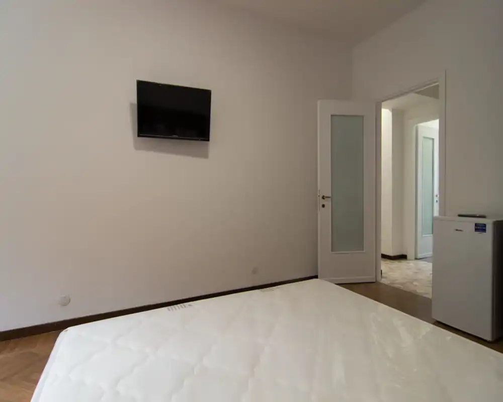 Camera da letto minimal con materasso in primo piano, TV a parete, pareti neutre e porta che conduce al corridoio. Spazio pulito e semplice adatto per allestimenti o annunci di affitto.