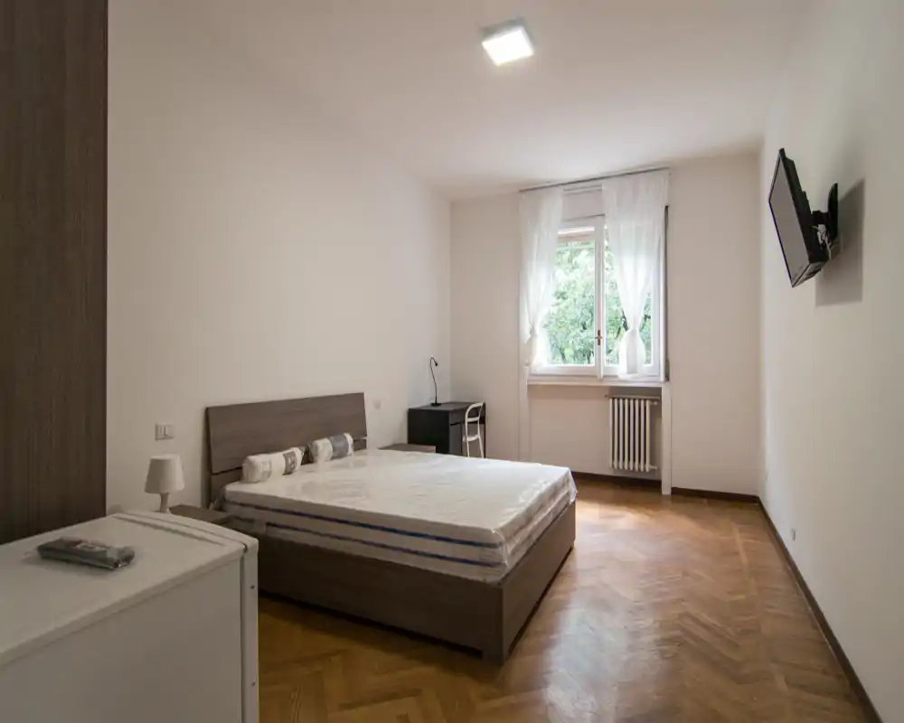 Camera da letto luminosa e arredata in modo essenziale con letto matrimoniale, pavimento in parquet e finestra che offre luce naturale. Spazio pulito e ordinato adatto a foto per affitto.