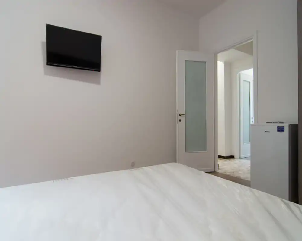 Camera da letto minimal e pulita con il letto rifatto in primo piano, una TV a parete e una porta aperta che conduce a un corridoio luminoso. L'ambiente è ordinato con finiture moderne e semplici.