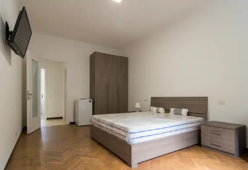 Dormitorio moderno y limpio con cama doble, cabecero y mesita a juego, armario y TV de pared; luminoso y de estilo minimalista, ideal para alquileres de corta estancia.