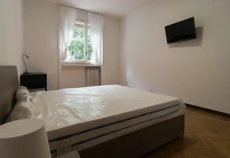 Dormitorio luminoso y sencillo con cama doble grande (colchón aún envuelto), ventana con cortinas traslúcidas y TV en la pared — decoración neutra adecuada para anuncios.