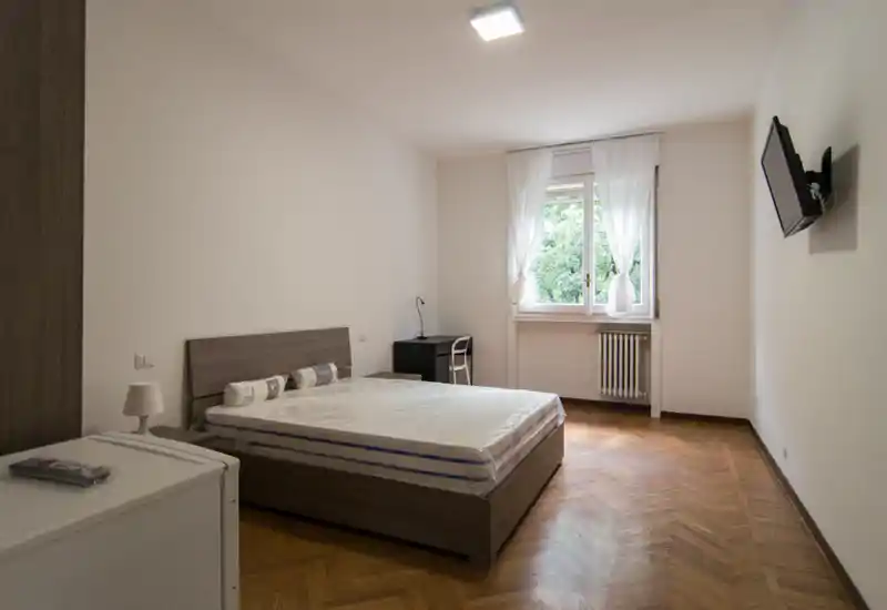 Dormitorio luminoso y minimalista con cama doble sobre estructura de madera, suelo de parquet y ventana que aporta luz natural. Escritorio sencillo y televisor mural completan la distribución funcional, ideal para anuncios.