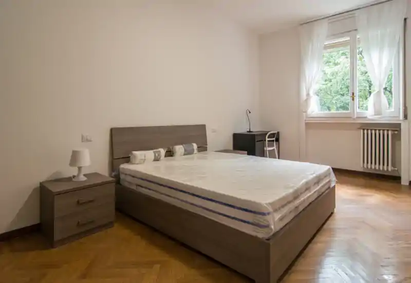 Dormitorio luminoso y de mobiliario sencillo con cama doble, mesita de noche y escritorio junto a la ventana; la luz natural y el parquet aportan calidez.