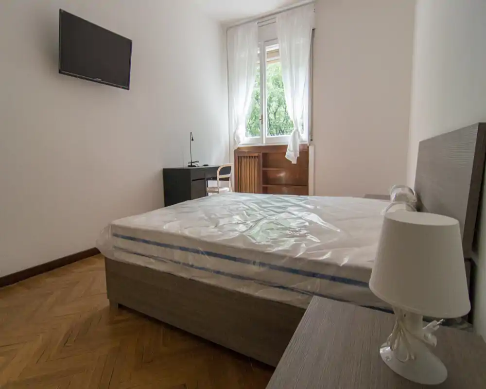 Camera da letto semplice e luminosa con letto matrimoniale (materasso ancora avvolto nella plastica), comodino con lampada, TV a parete e scrivania vicino alla finestra che offre luce naturale.