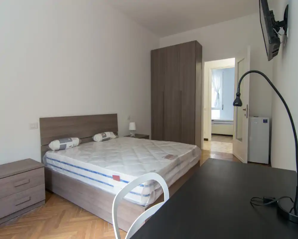 Camera da letto luminosa e minimalista con letto matrimoniale, comodini coordinati e armadio. Si notano il pavimento in parquet e una scrivania in primo piano.