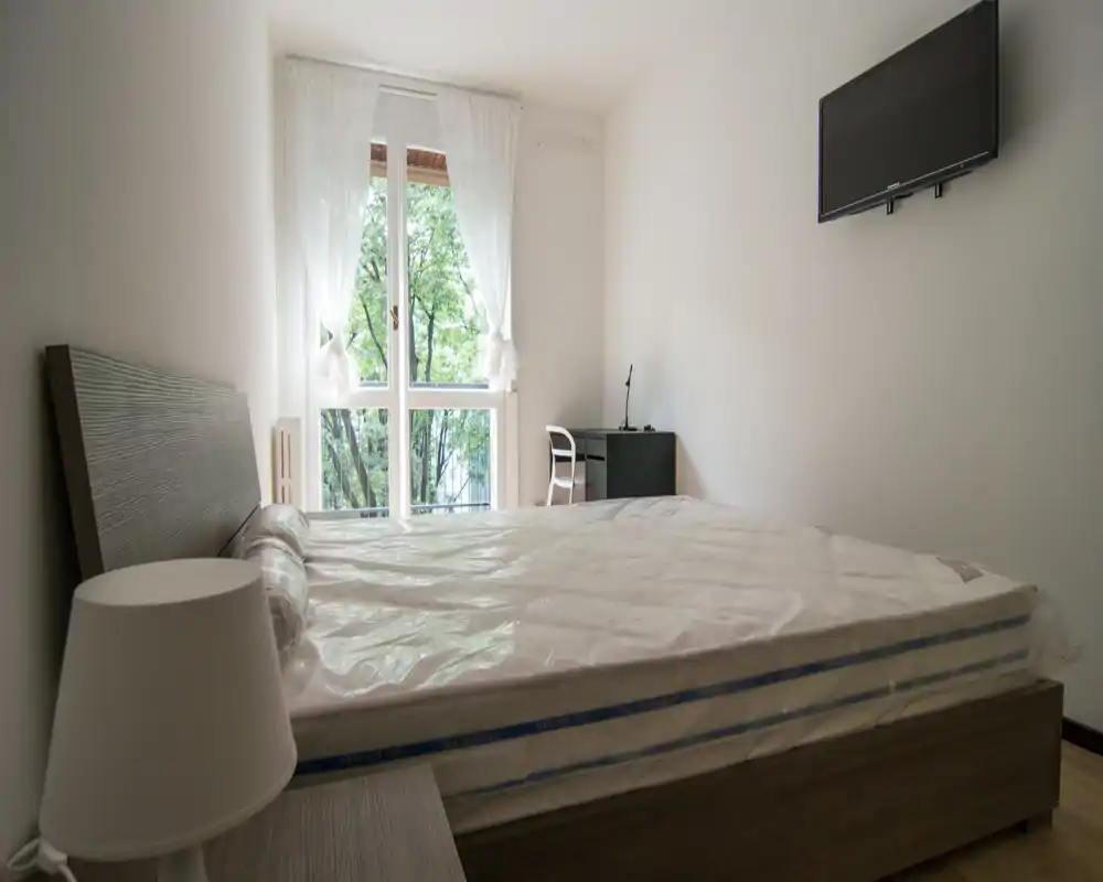 Dormitorio luminoso y de mobiliario sencillo con cama doble, lámpara de mesita y escritorio junto a la ventana. Ventanal con cortinas translúcidas que aporta luz natural y televisor de pared para un aspecto minimalista.