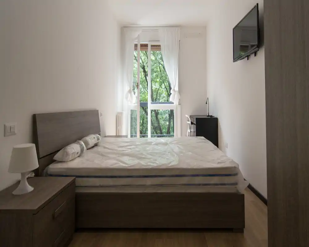 Dormitorio minimalista con cama doble, mesita de noche y luz natural procedente de una gran ventana; mobiliario de madera neutra y cortinas sencillas crean un espacio luminoso y tranquilo, ideal para alquileres.