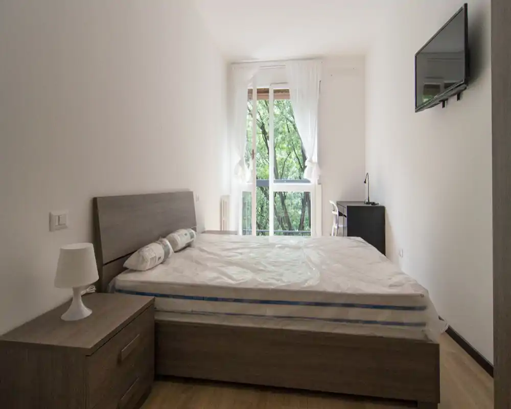 Dormitorio luminoso y minimalista con cama doble, mesita de noche y lámpara, televisor en la pared y escritorio junto a una gran ventana con vistas al verde.
