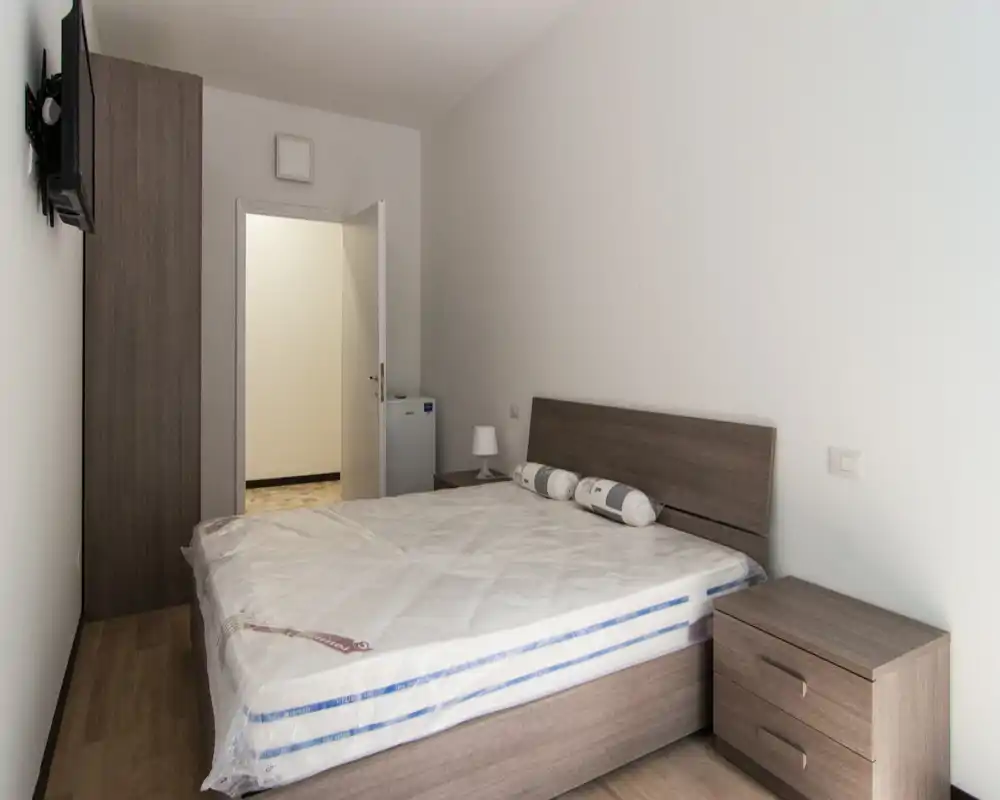 Dormitorio sencillo y bien encuadrado con una cama doble grande, cabecero y mesitas de noche de madera a juego. Tonos neutros y decoración mínima que crean un espacio limpio y funcional, ideal para alquileres temporales.