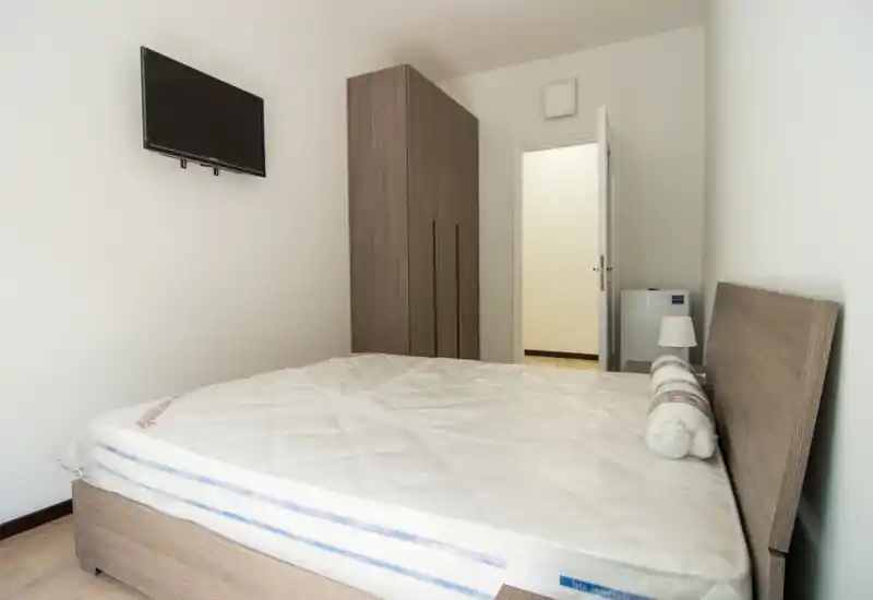 Camera da letto luminosa e minimal con letto matrimoniale, testata e armadio in legno; lampada da comodino e TV a parete per una sistemazione pratica.