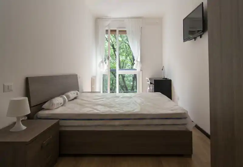 Camera da letto minimalista con letto matrimoniale, comodino e luce naturale da una grande finestra; mobili in legno neutro e tende semplici creano uno spazio luminoso e tranquillo, ideale per affitti.