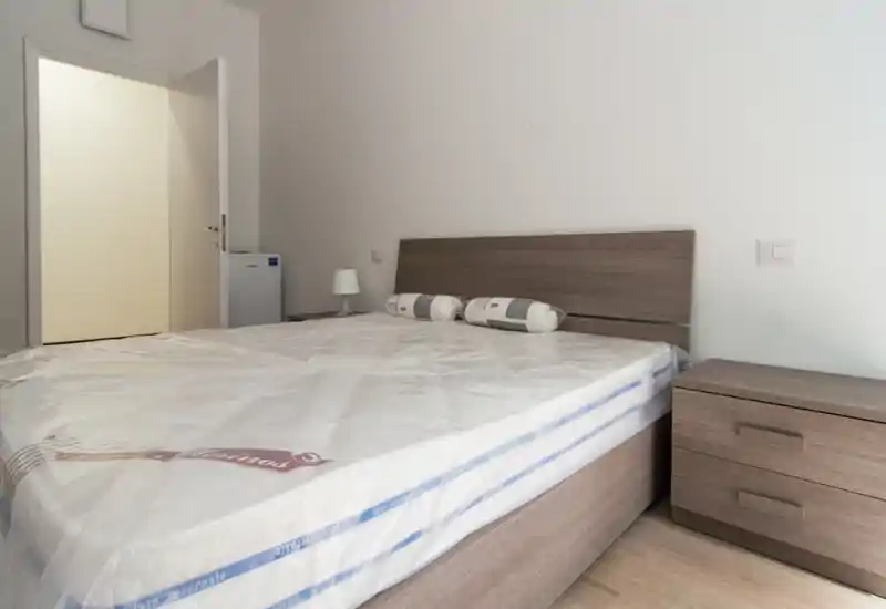 Camera da letto luminosa e moderna con grande rete e testiera in legno, comodino e lampada; materasso nuovo visibile completamente, stile pulito e minimale perfetto per annunci.
