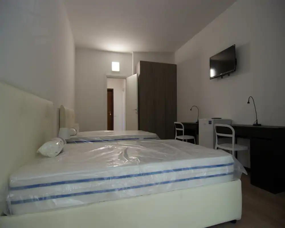 Bright, simply furnished bedroom with two single beds still in protective covers, a wardrobe and wall-mounted TV—ideal for short stays or guest rooms.