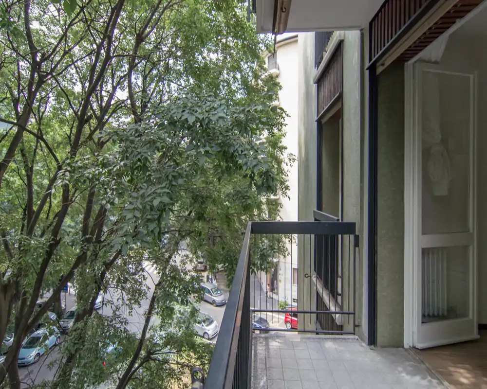 Sunny balcony overlooking a tree-lined street, with metal railing and tiled floor — ideal for morning coffee or light outdoor seating.