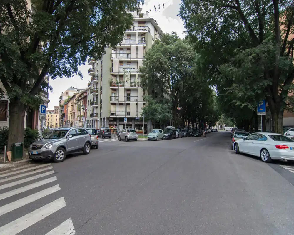 Strada urbana fiancheggiata da alberi con auto parcheggiate e palazzi residenziali di media altezza; inquadratura ampia ideale per mostrare il quartiere e la facciata stradale.