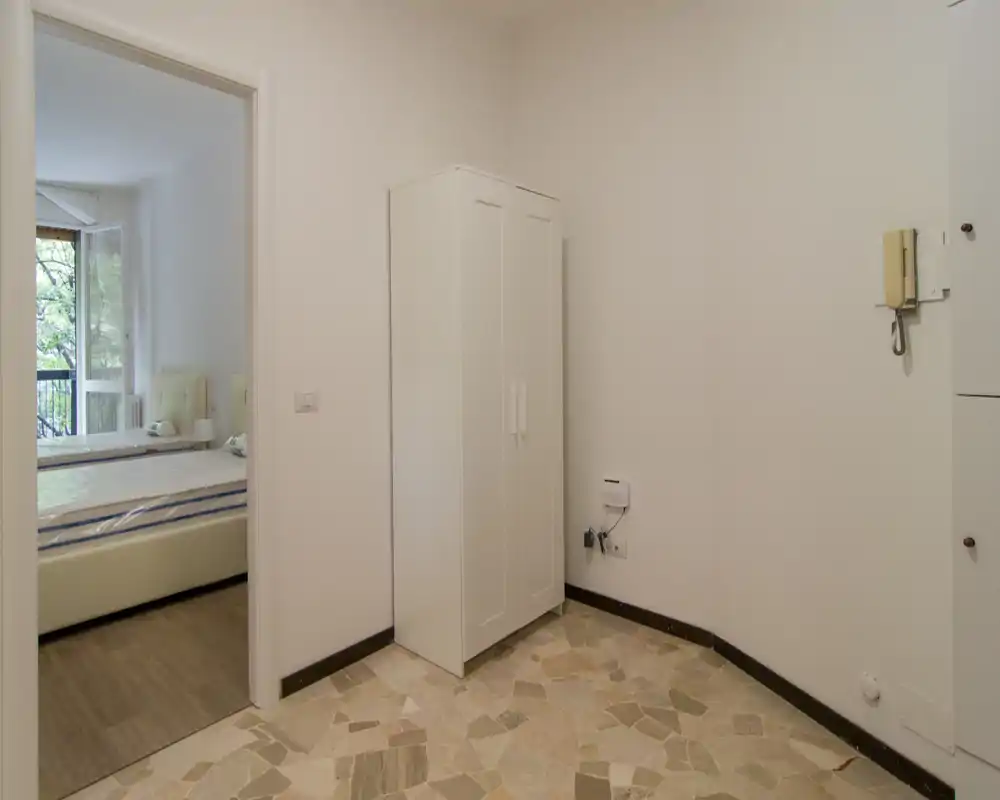 Piccolo ingresso con pavimento in cementine e un armadio bianco; attraverso la porta è visibile una camera da letto illuminata dalla finestra.