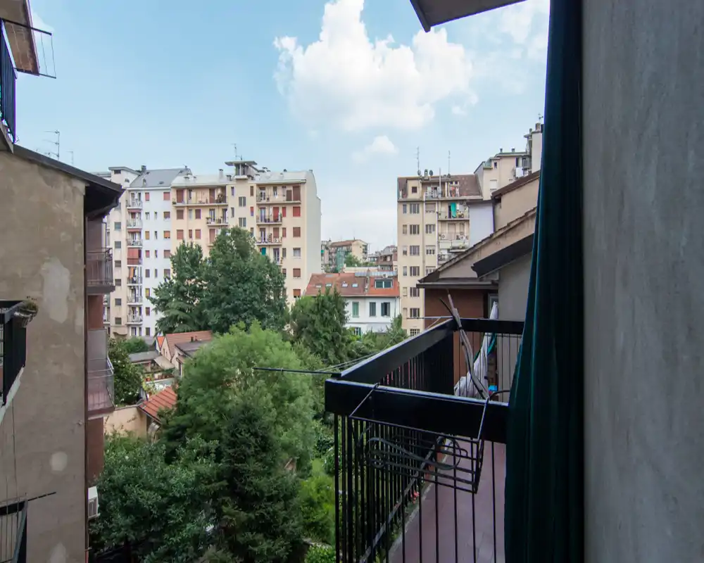 Balcone con vista su un cortile residenziale con alberi e palazzi circostanti, fotografato in pieno giorno.