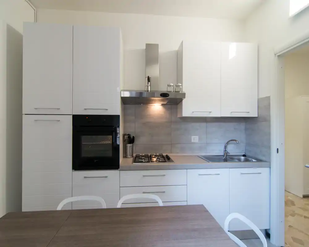 Cucina moderna e compatta con mobili bianchi, forno integrato, piano cottura a gas e lavello in acciaio; tavolo in legno in primo piano.