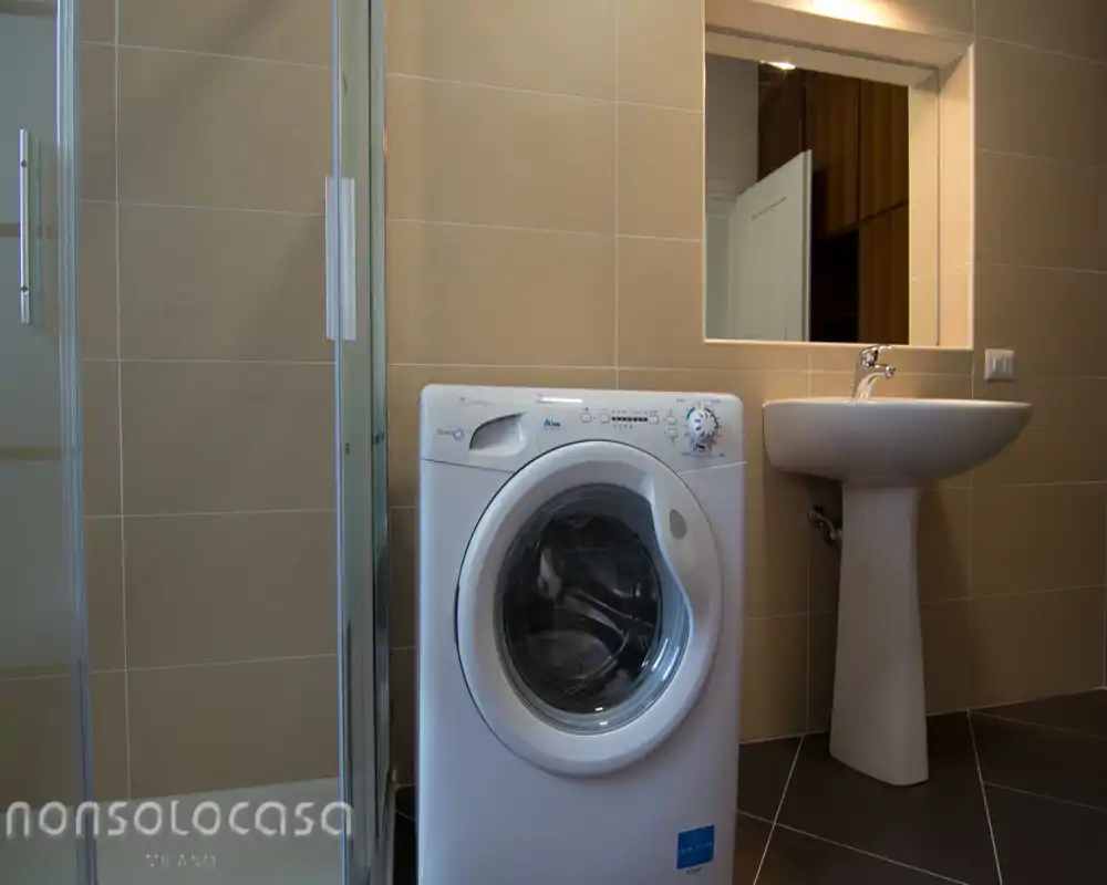 Bagno moderno compatto con pareti rivestite, lavabo a colonna, specchio e lavatrice; toni neutri e box doccia in vetro visibile a sinistra.