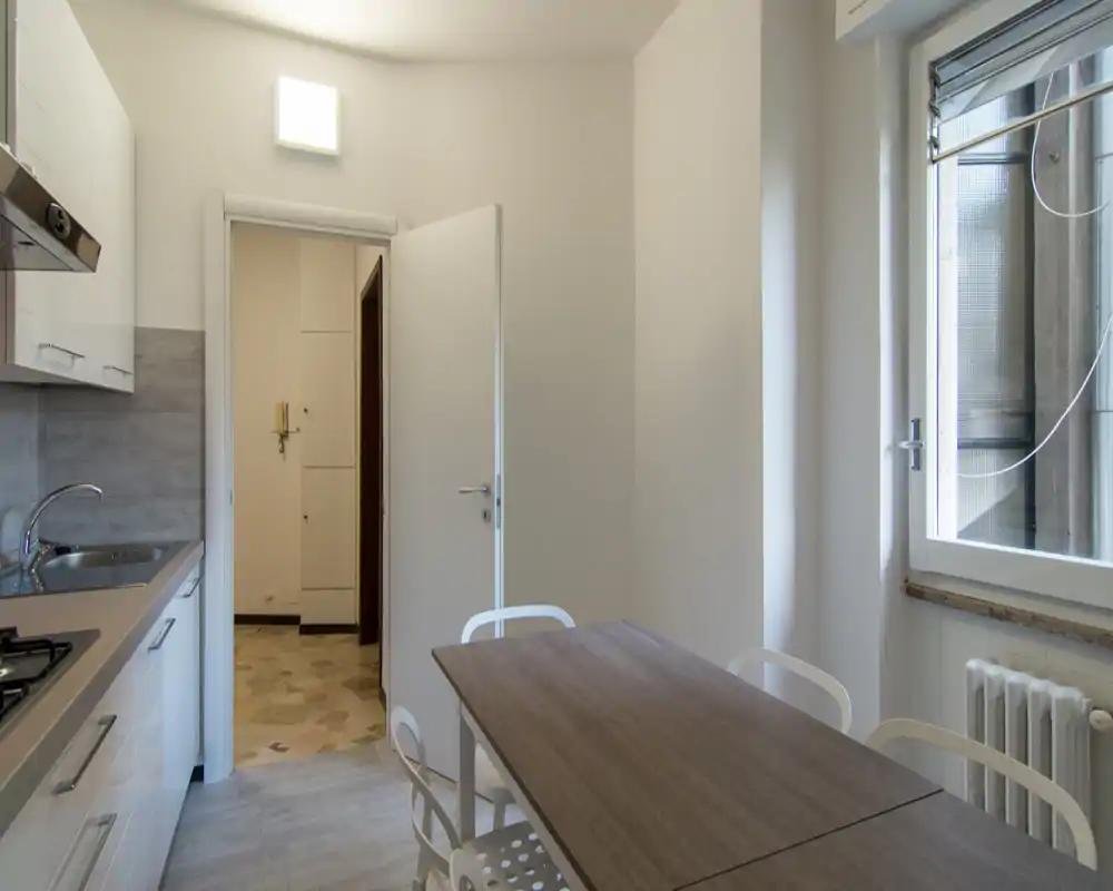 Cucina compatta e moderna con tavolo in legno, quattro sedie bianche e una finestra che offre luce naturale. Si vedono mobili puliti, lavabo e piano cottura, ideale per cucinare e pranzi informali.