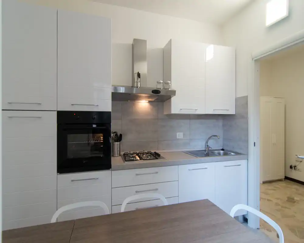 Cucina moderna con mobili bianchi, forno incassato, piano cottura a gas e lavello in acciaio. Il tavolo in primo piano suggerisce un’area pranzo informale; l’ambiente è luminoso e pulito.