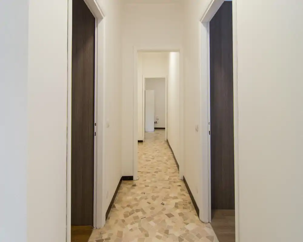 Corridoio lungo e luminoso con pavimento in piastrelle a motivo e pareti neutre, che mostra più porte e un percorso chiaro attraverso l’appartamento.