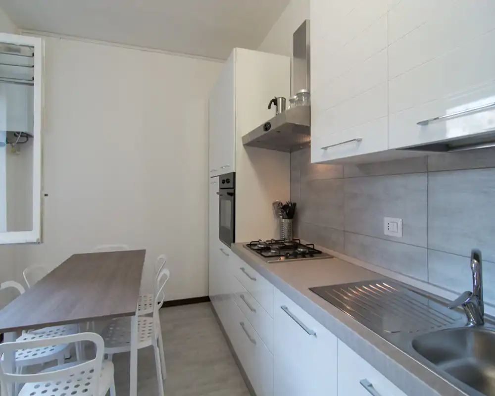 Cucina moderna e compatta con elettrodomestici integrati, piano cottura a gas, lavello in acciaio e un piccolo tavolo da pranzo con sedie — luminosa, pulita e funzionale.