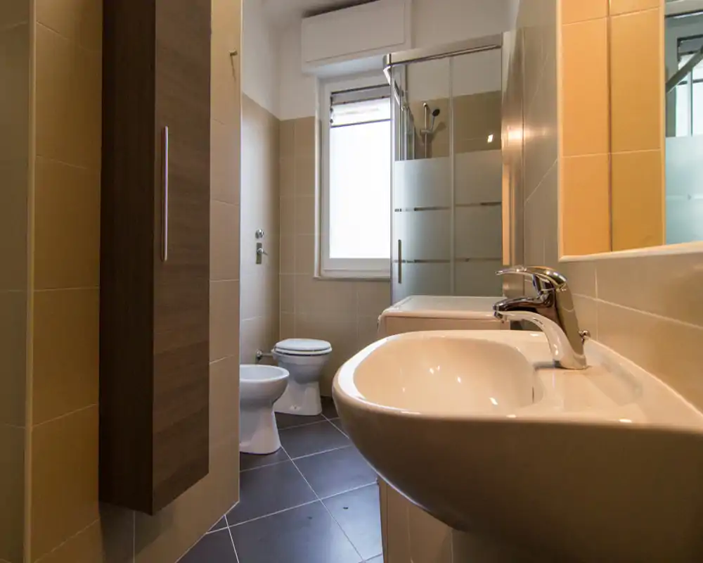 Bagno moderno e ben illuminato con lavabo in primo piano, bidet e WC vicino alla finestra e box doccia in vetro; piastrelle neutre e un armadietto a parete alto offrono spazio per riporre.