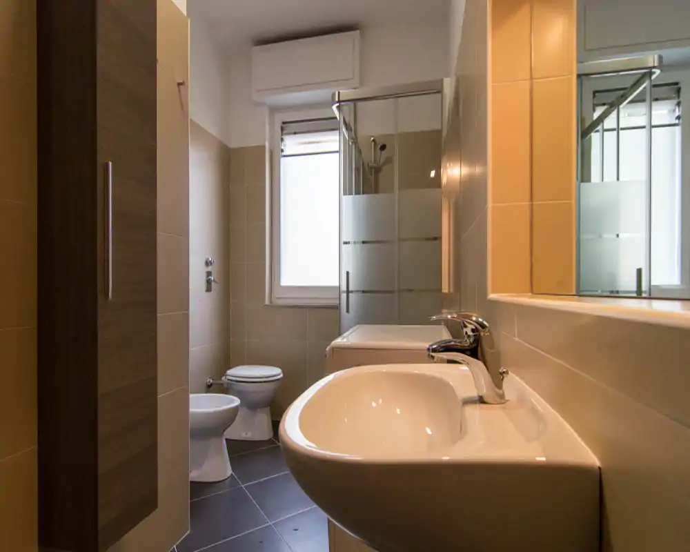 Bagno moderno e pulito con lavabo in primo piano, wc e bidet visibili e una doccia con box in vetro vicino alla finestra.