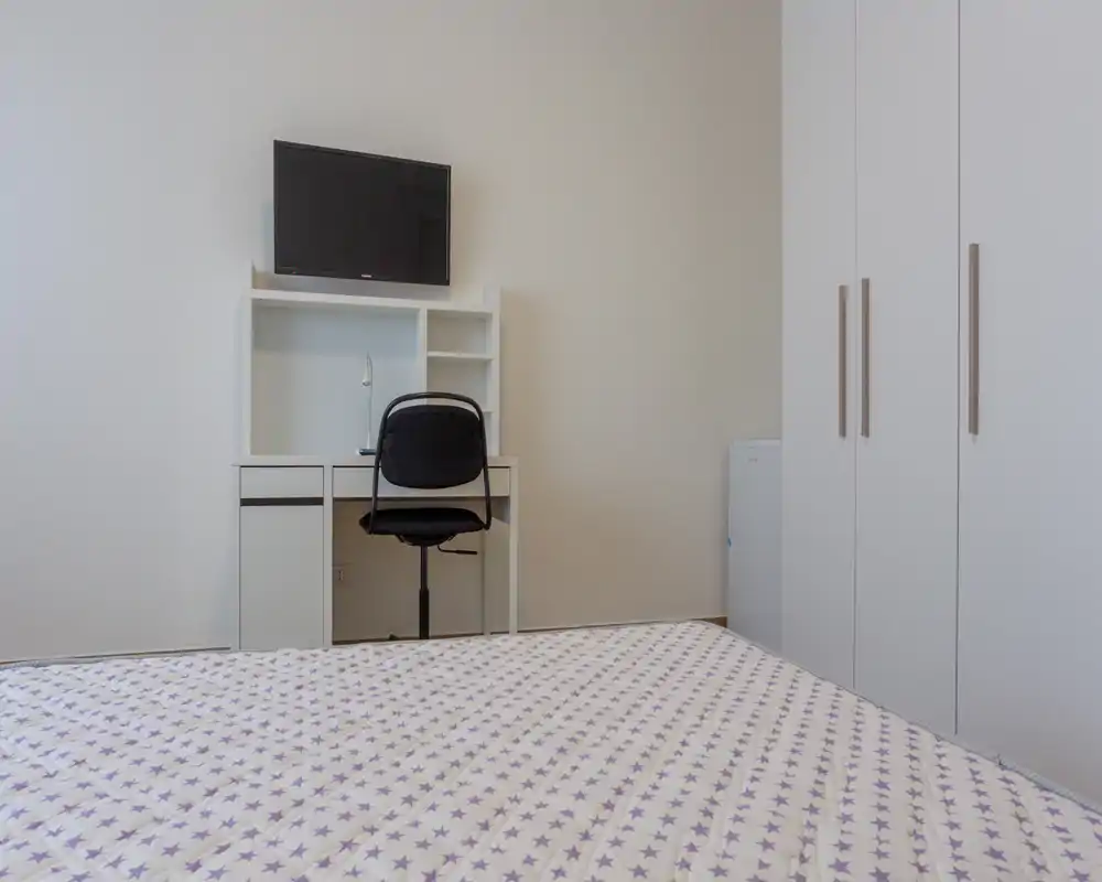 Dormitorio sencillo y ordenado con colcha estampada, escritorio blanco con estanterías, TV en la pared y armario — luminoso, minimalista y bien preparado para alquileres de corta estancia.