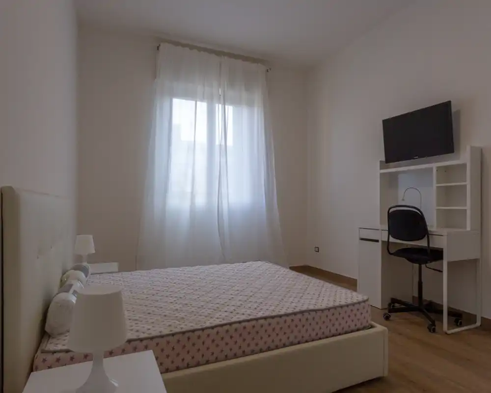 Dormitorio luminoso y de mobiliario sencillo con cama doble, lámparas de mesilla y un escritorio con TV en la pared. La luz natural filtrada por las cortinas aporta una atmósfera tranquila y neutra, ideal para alquileres de corta estancia.