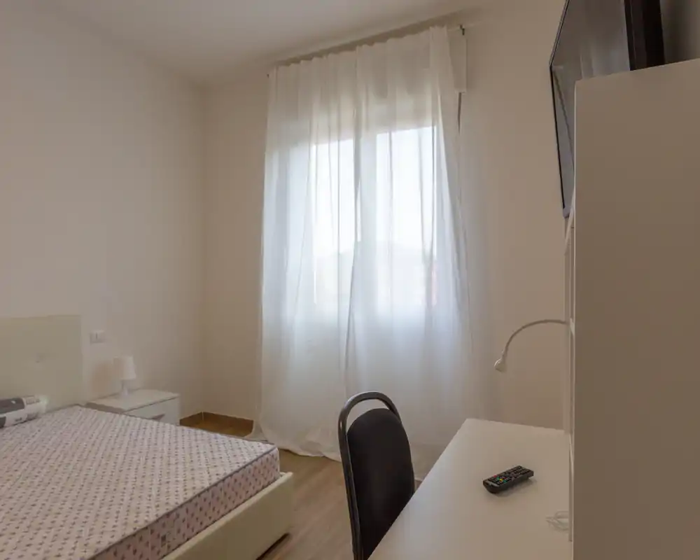 Dormitorio luminoso y minimalista con cama individual, mesita y escritorio frente a la ventana con cortinas translúcidas — líneas sencillas y luz natural.