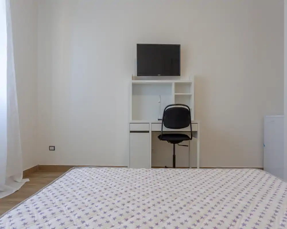Habitación minimalista con una cama grande en primer plano, escritorio, silla y TV en la pared al fondo. Espacio luminoso y sencillo, ideal para alquileres de corta estancia.