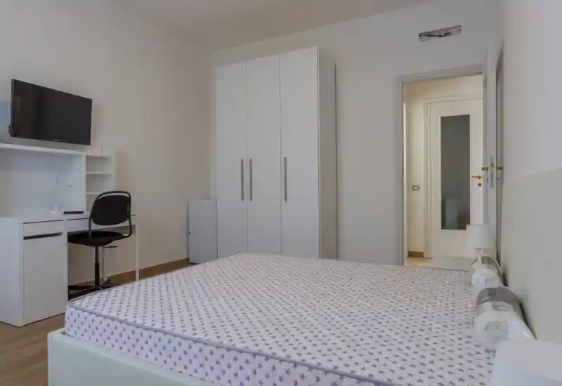 Dormitorio luminoso y sencillo con cama doble, lámpara de noche, armario y pequeño escritorio con TV — decoración neutra y limpia ideal para un anuncio de alquiler.